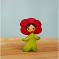 Figurine with red flower head and green body on a wooden surface with light blue background