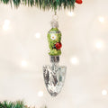 A Christmas tree ornament shaped like a garden trowel with a green handle decorated with white flowers and a small red ladybug, hanging from a tree branch with lights in the background.