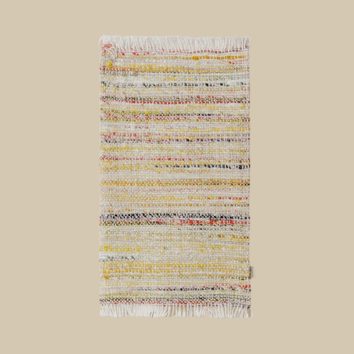 A rectangular woven rug with fringed edges, featuring horizontal stripes of white, yellow, red, and blue yarn on a beige background, shown against a solid beige backdrop.