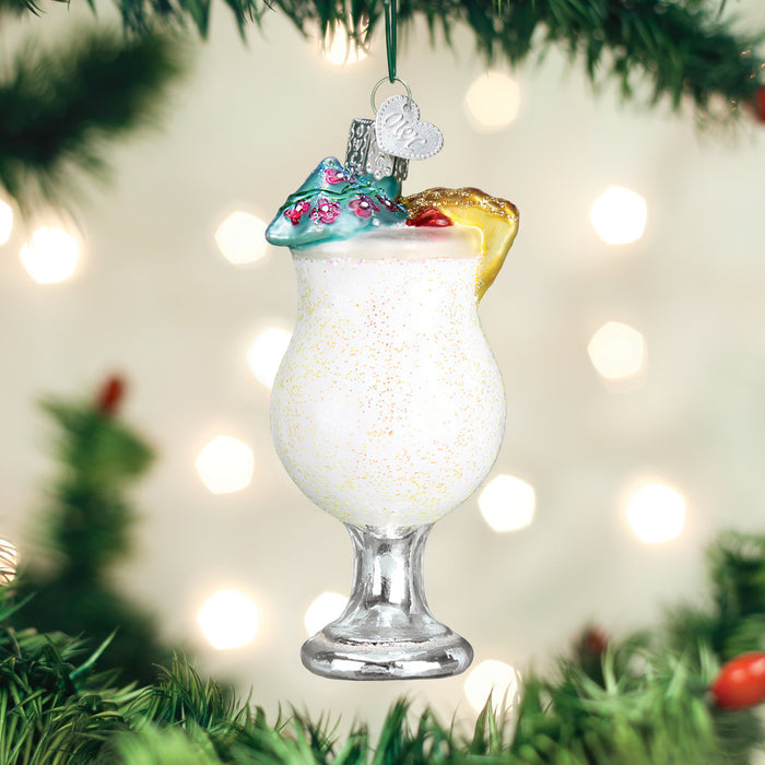 A decorative ornament shaped like a tropical cocktail with a slice of pineapple, cherry, and umbrella, hangs on a Christmas tree with green branches and blurred white lights in the background.