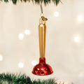 A Christmas tree ornament shaped like a plunger, with a gold handle and a shiny red base, hangs from a green tree branch against a blurred background of white lights.