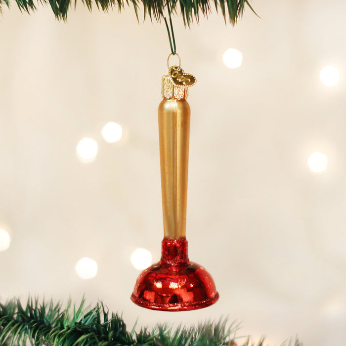 A Christmas tree ornament shaped like a plunger, with a gold handle and a shiny red base, hangs from a green tree branch against a blurred background of white lights.
